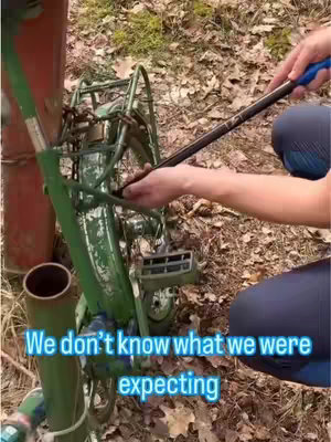 A close-up shot shows a person's hand using a crowbar to try to unlock a chain that secures an old green bicycle to a metal pole. The scene is outdoors, and the ground is covered in leaves.