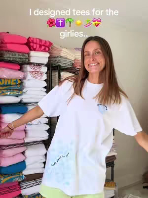 A woman stands smiling in front of shelves stacked with folded t-shirts. She wears a white t-shirt with a blue design and short yellow-green shorts. The text "I designed tees for the girlies.." with associated emojis is overlaid at the top.