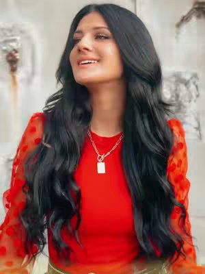 A woman with long, dark hair and a red top is smiling and looking upwards. She wears a gold chain necklace. The background is a blurred, neutral color.
