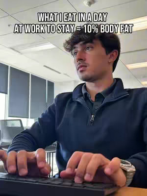 A medium shot of a man sitting at a desk, typing on a keyboard. He is wearing a dark blue fleece jacket, with a watch on his left wrist. The background shows an office setting with large windows and blinds. Text overlayed across the top reads 'WHAT I EAT IN A DAY AT WORK TO STAY ≈ 10% BODY FAT.'
