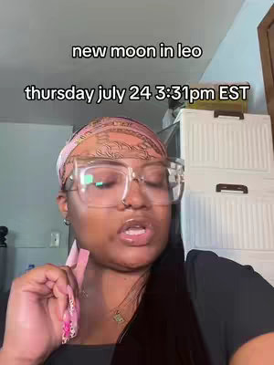 The image is a selfie-style shot of a person, likely taken indoors. The person is positioned in the foreground with a background that appears to be a room, though its details are slightly blurred. The individual is the main focus, and they have a headscarf. They are wearing large, clear-framed glasses that reflect light. The person's mouth is slightly open, and they appear to be speaking or expressing something. Above the person, there is text with information about "new moon in leo" and the date and time.