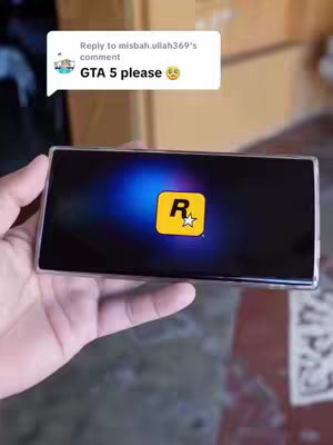 A hand holding a black smartphone displaying the Rockstar Games logo from Grand Theft Auto 5. A comment bubble appears in the top left corner. The background seems to be a wall.