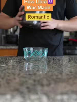 A person is shown from the torso up, holding a dark container above a clear, patterned glass bowl on a speckled countertop. The background is a kitchen setting with a blurred view of cabinets and appliances. Text overlays in bright colors are present on the top of the image