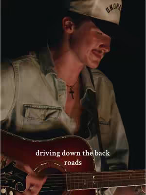 A man wearing a baseball cap and a denim shirt is playing a guitar. Text overlay reads "driving down the back roads". The lighting appears to be low, creating a moody atmosphere.