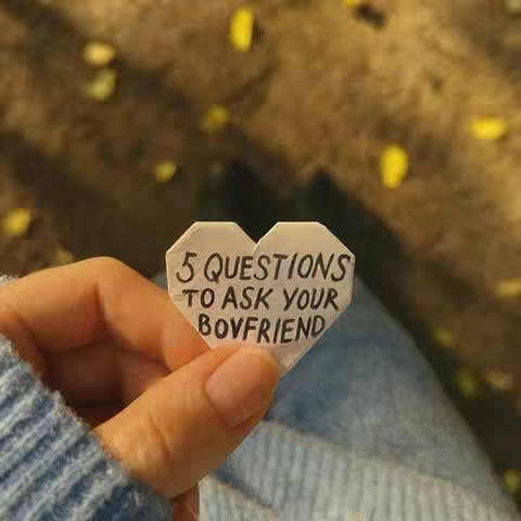 A hand holding a paper heart with text, set against a blurred background of fall leaves and a person wearing jeans and a blue sweater. The composition focuses on the heart and hand, suggesting a message about love and relationships.