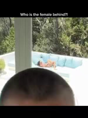 The image shows a close-up of a person's head, with the top of their head filling the lower portion of the frame. Above them, a blurred scene depicts an outdoor setting with greenery and a person lying down on a cushioned surface. A text overlay at the top reads 'Who is the female behind?!' in a bold, white font.