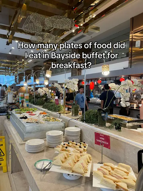 An interior shot of a buffet area in a restaurant, with various food and beverage stations visible. The foreground includes trays of sandwiches and plates stacked up. There are multiple people at different stations. The overall scene gives an impression of abundance and variety.