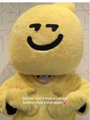 The cover image shows a person in a full-body yellow Labubu costume. The figure's face is a large, round, and yellow head with a black smiley face and black eye-shaped markings above. Below the head is a peek-a-boo detail of a grey figure. Overlaid on the image is the text: "Sue me but I'd trust a Labubu before I trust a man again".