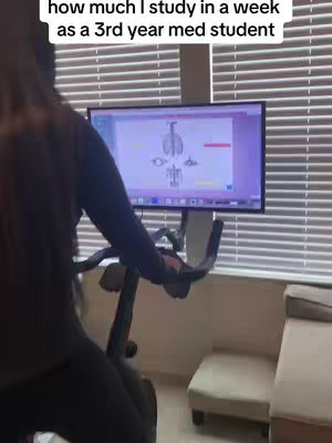 A person is shown from the back, their torso partially visible, as they ride a stationary exercise bike. A screen displaying a medical diagram is positioned in front of them. The background includes window blinds and a cream-colored armchair, suggesting an at-home setting.