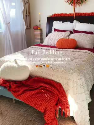 A cozy bedroom scene featuring a bed with white bedding and a red blanket draped over the foot. An orange pumpkin-shaped pillow sits on the bed. The room has a window with white curtains on the left, and a wooden decorative piece on the right, enhancing the fall theme.