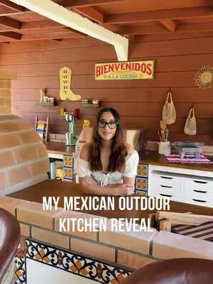 A woman with glasses smiles inside a partially constructed outdoor kitchen. The scene is warm-toned, with a brick oven and wooden structures. Overlay text reads 'MY MEXICAN OUTDOOR KITCHEN REVEAL' 