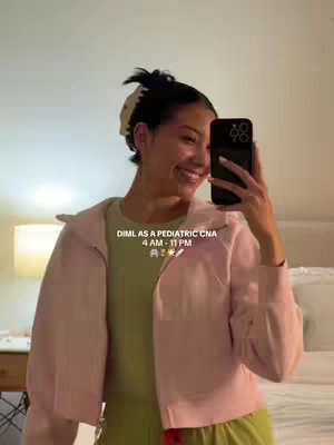 A woman is taking a selfie, holding a phone in front of her face. She is wearing a light pink jacket over a light green top, complemented by wide-legged, light green pants. Her hair is pulled back, and she is smiling at the camera. The background is softly lit with a lamp and a bed visible. Text overlay states 'DIML AS A PEDIATRIC CNA' and the work hours along with some emojis.