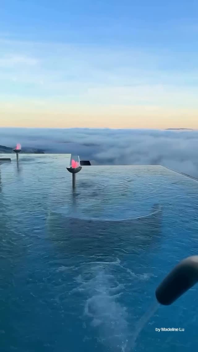 The cover image shows an infinity pool. The blue water of the pool merges seamlessly with the horizon, creating the illusion of swimming among the clouds. Two lit, slender torches are placed in the pool. A person's arm and hand are visible in the lower right corner, with the text "by Madeline Lu" at the bottom.