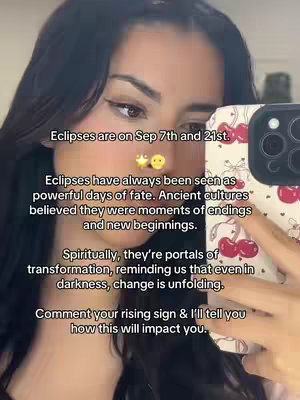 A selfie-style video with a female creator holding up a cherry-patterned phone. Text overlays provide information about eclipses, their significance, and call to action for viewers to comment their rising sign. The background is a light-colored room, with the creator facing the camera directly.