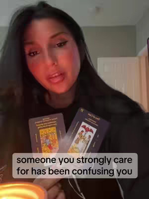 A medium shot of a woman with dark hair and makeup, holding tarot cards. She appears to be speaking, with text overlayed at the bottom of the image.