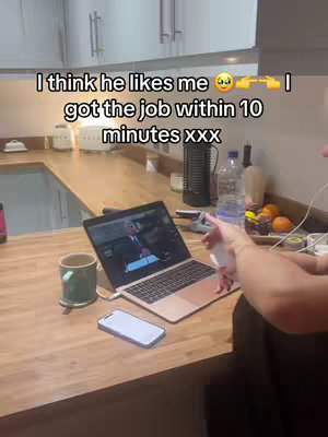 A person is sitting at a wooden counter in a kitchen with a laptop, a phone, and a mug. The laptop screen shows a man speaking, likely a video call. Beside the laptop is a mug. The person's arm and part of their body are visible, as is a water bottle and some oranges in the background of the kitchen. Text on the top overlay says 'I think he likes me' with an emoji, then 'I got the job within 10 minutes xxx'.