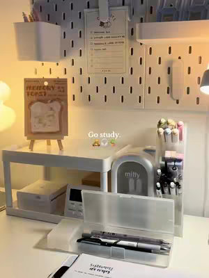 A well-lit, tidy desk setup with various study-related items. A white pegboard with small holes is mounted on the wall, holding various stationery items in white holders. A white shelf with a white stand with notes, pens, and various containers. A clear plastic container filled with pens sits on the desk. A small, grey 'miffy' object is placed. The desk shows a clean, organized aesthetic.