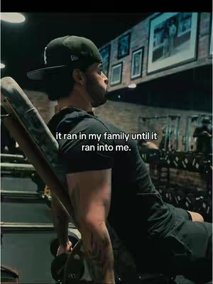 A man in a black cap and shirt is mid-workout in a gym. He is leaning back on a weight bench, gripping a dumbbell, with a focused expression. Text overlayed mentions family and running into him. The background shows gym equipment, framed pictures on the wall, and a brick wall.