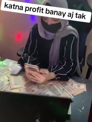 A woman wearing a hijab and a face covering is seated at a table with what appears to be a large amount of cash spread out across the surface. She is holding a phone and looking at it. The background is dimly lit with blurred colors.