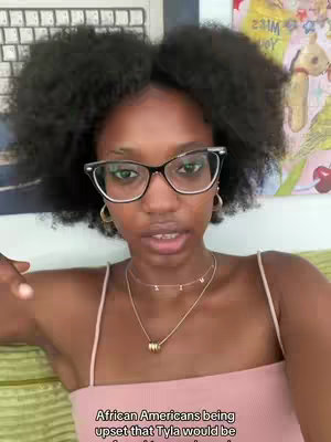 A selfie-style video features a young woman with dark curly hair, clear glasses, and multiple necklaces against a backdrop that is partially visible. She appears to be speaking directly to the camera.
