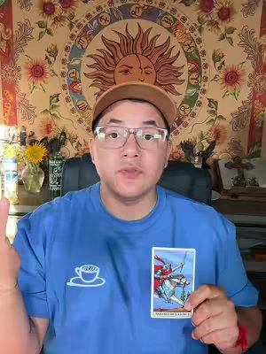 A man with glasses and a hat is holding a tarot card in front of his chest. The background is a tapestry with astrological symbols and the sun. The man wears a blue shirt and the overall tone is warm.