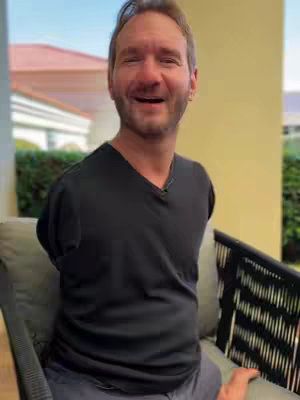 A medium shot depicts a smiling, light-skinned man seated in a chair on what appears to be an outdoor patio. He has short, light brown hair and a neatly trimmed beard. He is wearing a black V-neck shirt. The man is seated in a chair with a gray cushion, and his arms are missing from the image, visible only to about the elbows. The background is blurred, showing a glimpse of a roof, greenery, and a light-colored building. The overall impression is casual and friendly.