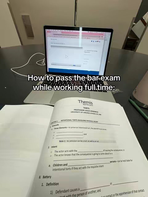 A well-lit overhead shot shows a laptop displaying a video screen, an open textbook on a desk and a pen. The laptop screen is open to a video player interface. The open book shows handwritten notes and text from a law textbook with the title "Torts". A pen rests on the right. The background suggests an office or study space.