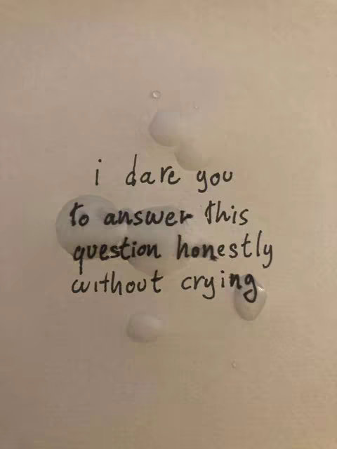 A close-up image on a textured, off-white surface, possibly a wall or a paper. Handwritten black text is centered, with the words arranged in a slightly curving design. The text says, 'i dare you to answer this question honestly without crying'. There are a few water droplets scattered on the surface, adding a textural element to the image.