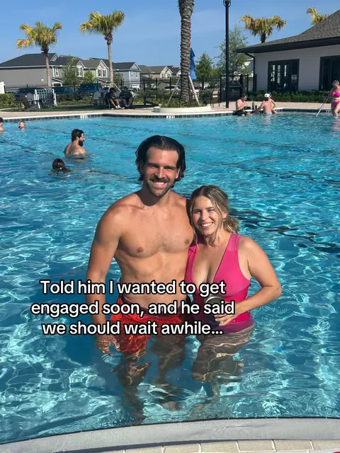 A couple stands in a swimming pool, smiling at the camera. The water is blue and reflects the sunny sky. The man is shirtless, while the woman wears a pink swimsuit. Behind them, other people swim in the pool, and buildings and palm trees are visible in the background.