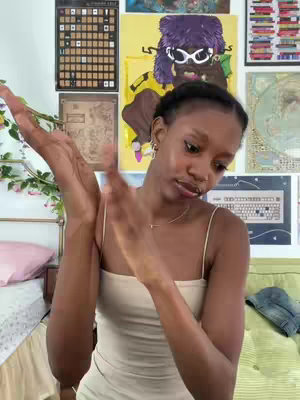 A young woman with dark skin and braided hair is clapping her hands in front of her. She is wearing a cream-colored spaghetti strap top and a delicate necklace. Her facial expression implies a lighthearted or appreciative mood. Her surroundings include room decor in the background.