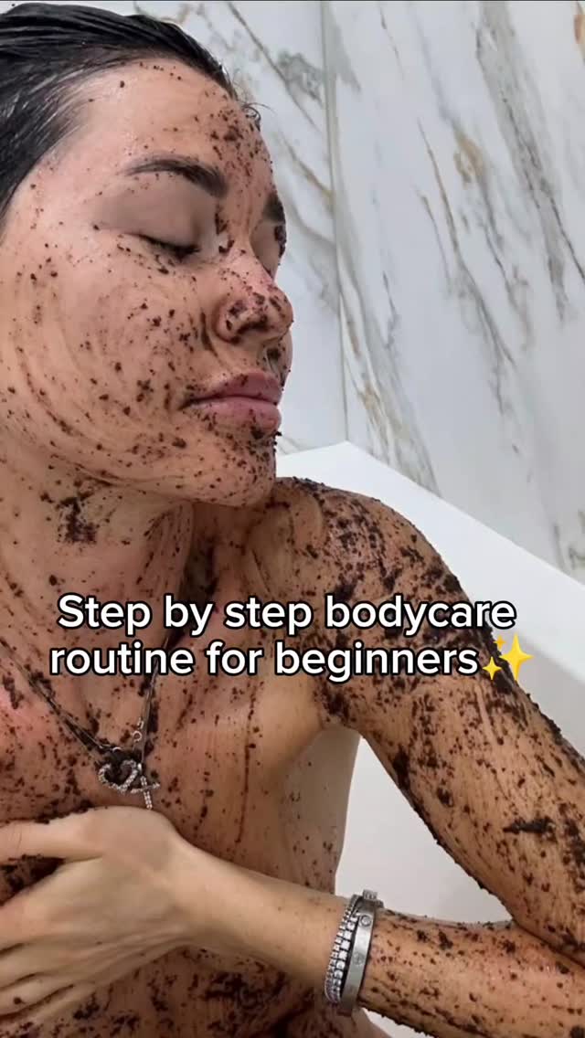 A woman covered in a dark, granular substance, possibly a body scrub, stands in a white tiled shower. The image focuses on her torso and face, with her eyes closed in apparent relaxation. Text is overlaid at the bottom, and overall composition is naturalistic with a focus on skincare.