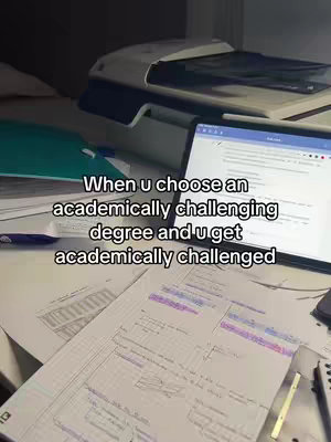 A well-lit, slightly angled shot of a desk covered in study materials. In the background, a printer sits on the desk and a laptop is open, displaying text on its screen. The desk is filled with scattered papers, notebooks, and a pen along with a water bottle.