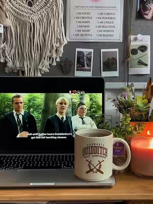 A cozy, indoor scene with a laptop displaying a scene from 'Harry Potter,' a mug with 'QUIDDITCH' branding, and a lit candle creates a warm, inviting atmosphere. The room is decorated with a macrame wall hanging, motivational prints, and potted greenery.