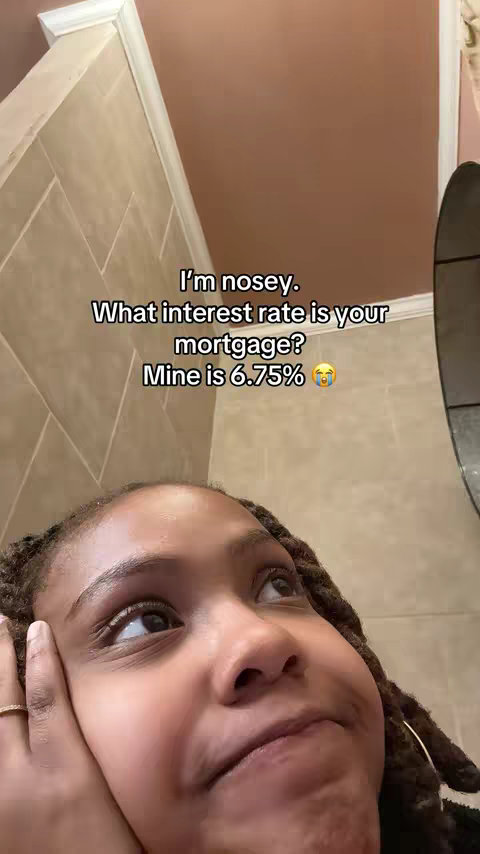 A close-up selfie of a young Black woman looking upwards and to the right. She has locs, and her right hand is resting on her cheek. The background is a bathroom with beige tiles and a brown ceiling.