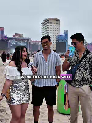 A casual shot at an outdoor event, featuring three people in the foreground. One person on the left is a woman holding a microphone, likely interviewing two men on the right. The setting includes a high-rise building, festival stages, and crowded environment behind.