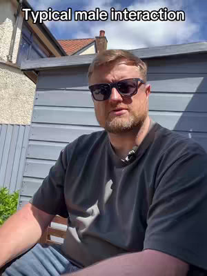 A man wearing sunglasses and a black t-shirt is sitting outside, with a shed and some greenery in the background. Text overlay says 'Typical male interaction'.