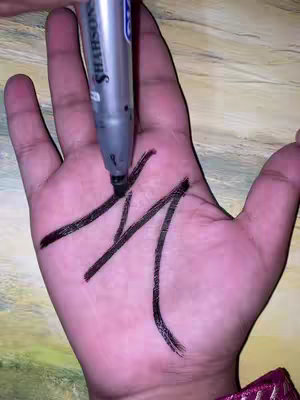 A hand with a dark, thick black marker drawing an 'M' shape on the palm. The hand is fair-skinned, and the background is blurred, with hints of a wooden surface and an indistinct colorful item at the bottom.