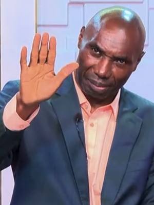 A medium shot featuring a middle-aged Black man with a bald head, wearing a teal suit jacket over a peach-colored dress shirt. He is holding his right hand up in a stopping gesture, with the palm facing toward the camera. The man has a stern expression on his face. The main source of light appears to be in front of the man and to his left.