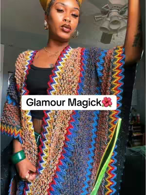 A woman with dark skin wearing a colorful patterned shawl over a black top, with a yellow head covering. A hook text overlay reads 'Glamour Magick' with a flower icon. The composition is a mid-shot with a room and fan in the background.