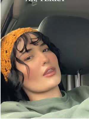 A woman facing the camera in a car, wearing an orange crocheted head covering and a green top. Her face is illuminated with makeup, and she looks contemplative.
