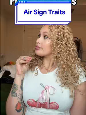 A woman with curly blonde hair is looking off to the side. She is wearing a white t-shirt with a cherry graphic. A text overlay titled "Air Sign Traits" is in the top left corner. Part of a room, including a bed and clothes, is slightly visible in the background.