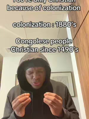 The image shows a man in a black hoodie with the hood up. He is looking down with a slightly concerned expression, and his hands are clasped together near his chest. The background appears to be an interior setting, possibly an office or living room, with a framed piece of art visible. Text overlays with white font on the top area of cover image that states, "You're only Christian because of colonization".