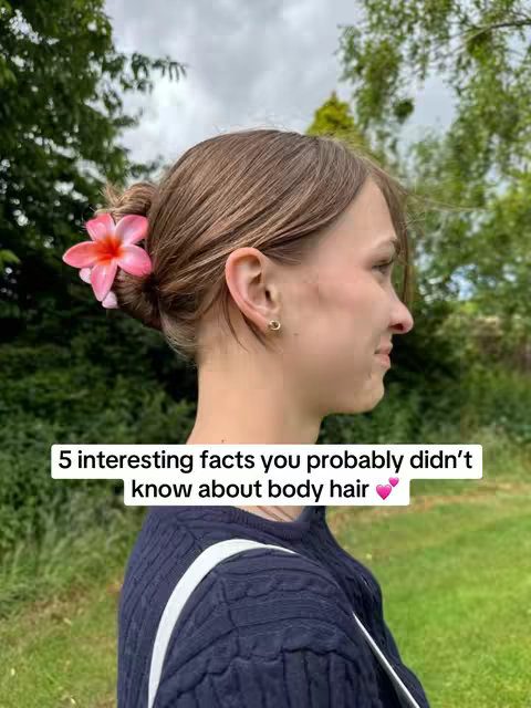 A woman with light brown hair styled in a bun is seen in a side profile view. She is wearing a dark blue sweater and a pink flower is tucked into her hair. Text on the image reads, "5 interesting facts you probably didn't know about body hair." The background is blurry with green trees and a cloudy sky.