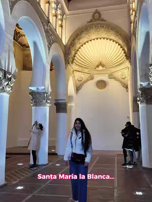 A woman stands in a large, ornate building with tall white arches and columns. The building has a high ceiling and detailed decorative elements. The woman is the central focus, and there are other people in the background.