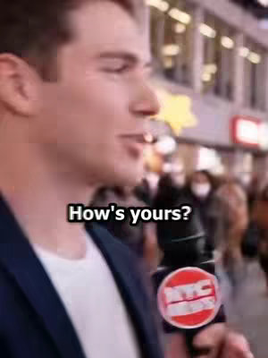A man wearing a navy blazer and a white undershirt speaks into a microphone with an "NYC NEWS" logo on it. He is in a crowded street setting with blurred background details, including lit buildings and a few masked individuals. The text overlay reads "How's yours?" in a black font.