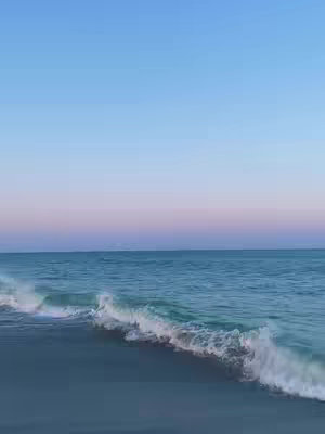 The image shows a serene beach scene at dawn or dusk. The sky is a gradient of blues and pinks, suggesting either sunrise or sunset. Below the sky, there is a vast expanse of water, its surface slightly rippled. In the foreground, waves gently roll onto a sandy beach. The scene has a calm and peaceful atmosphere, with soft light and colors.
