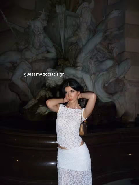 A woman poses against a backdrop of a large, ornate fountain with sculptures; she is dressed in a white lace outfit, with her arms raised and hands touching her hair; the text overlay reads "guess my zodiac sign".