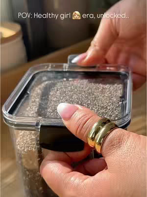 Close-up shot of a person's hand holding a clear container filled with small, dark seeds resembling chia seeds. The person is wearing a gold ring on their thumb and has polished fingernails. The background is slightly blurred and includes other containers and wooden surfaces, suggesting a kitchen setting.
