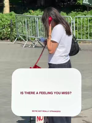 A woman stands on a sidewalk, holding a red telephone to her ear. A large, white and rectangular sign is in front of her, with the text 'Is there a feeling you miss?' in maroon. The background shows greenery, a metal fence, and general outdoor scenery, indicating a sunny day.