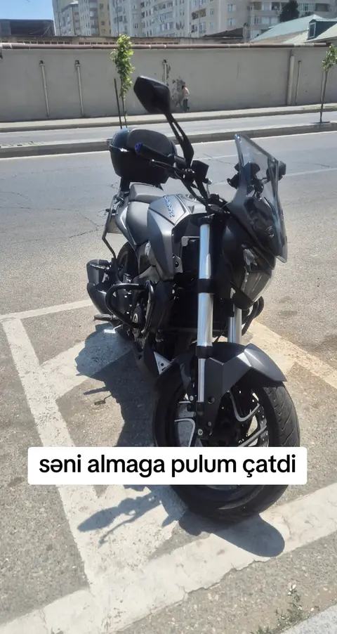 A black and gray motorcycle is parked on a paved area. The foreground shows parts of the motorcycle, including the front tire and fairing, and the background features a building and a clear sky. The word overlay in white is set in a rectangle.